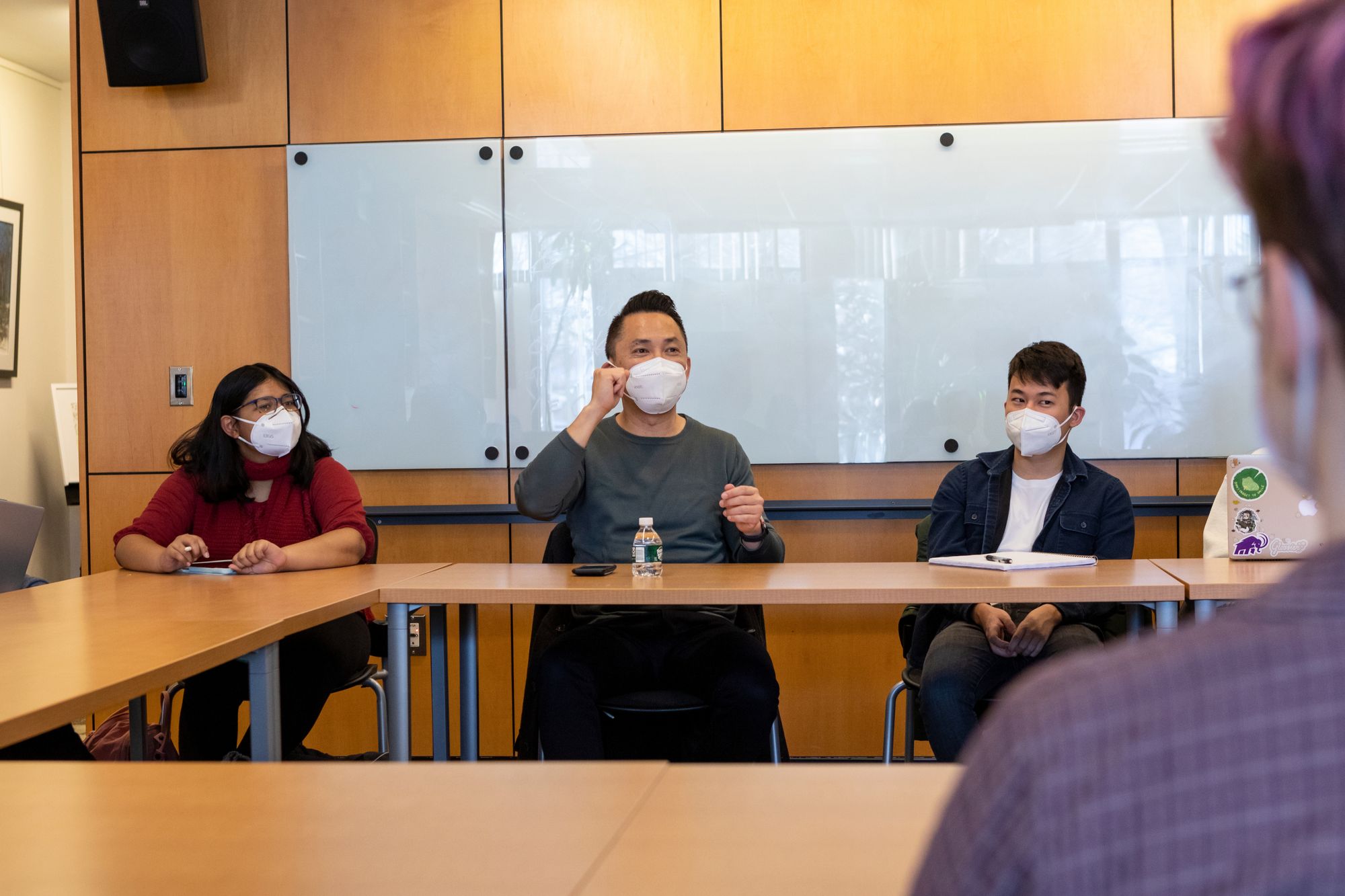 The image depicts Viet Thanh Nguyen animatedly speaking, sitting at a table between two students attending the talk.