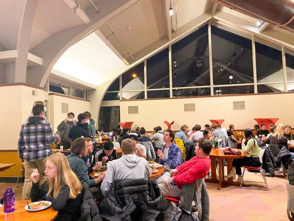 The image depicts a room crowded with students eating at tables in the back room of the dining hall.