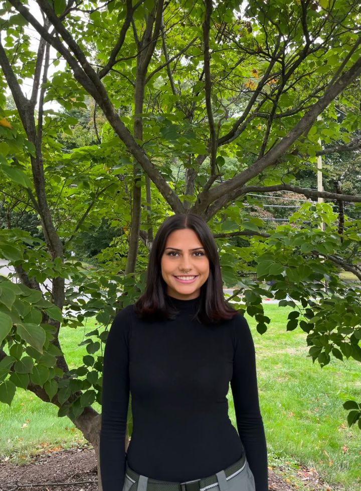 Renee, smiling and wearing a black mockneck, stands in front of a green tree.